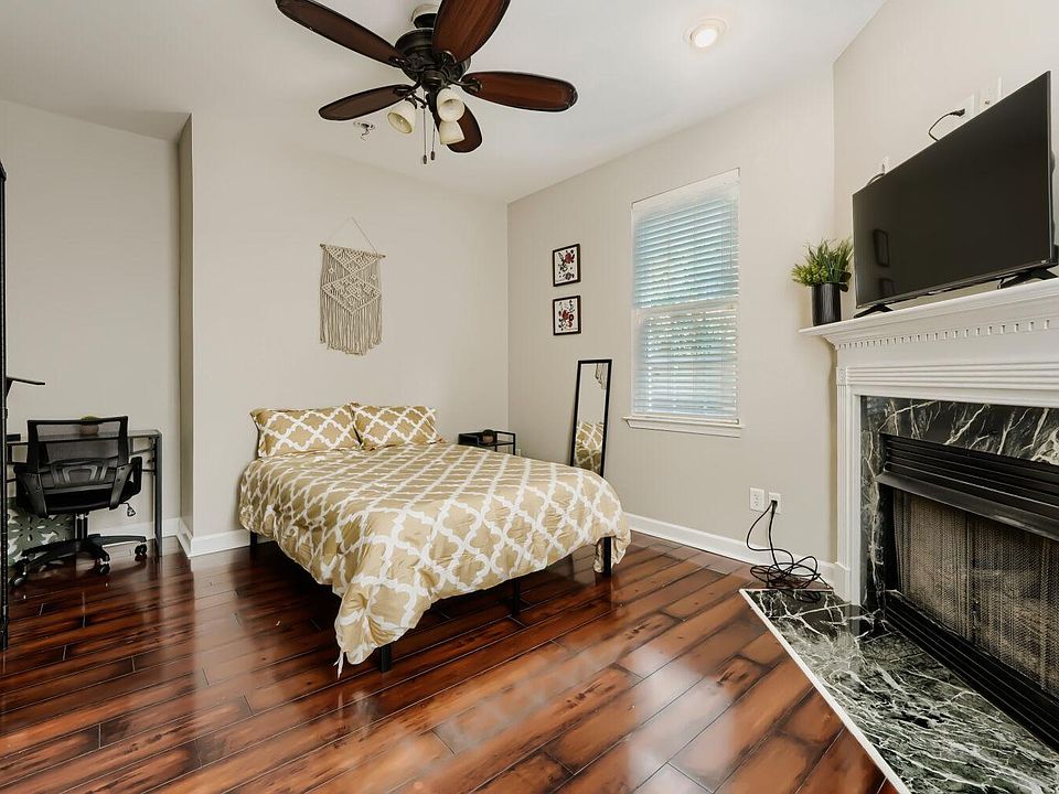 Spacious bedroom with hardwood floors, ceiling fan, desk area, and a decorative fireplace topped with a mounted TV.