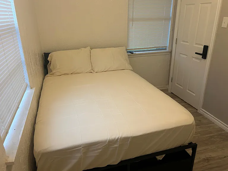 Furnished bedroom with a full-size bed, window blinds for privacy, and easy access to the hallway/entry.