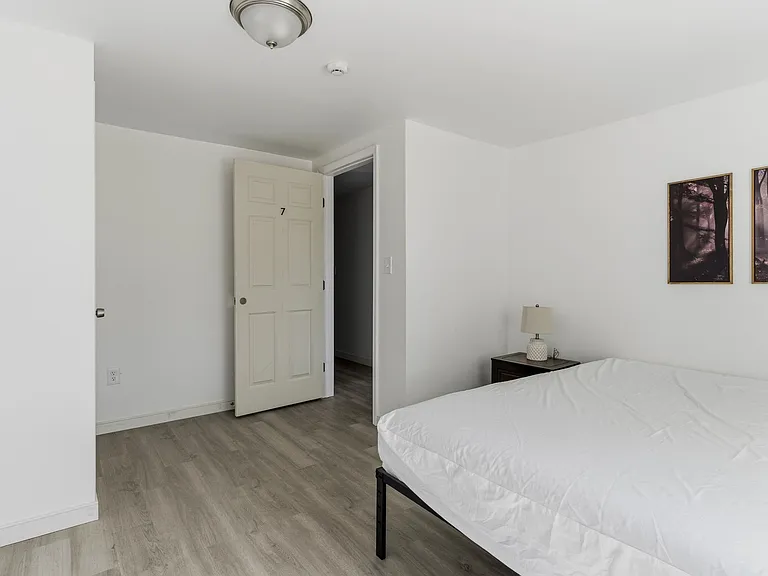 Simple furnished bedroom with bed, nightstand and lamp, durable wood-look flooring, and convenient access to the hall.