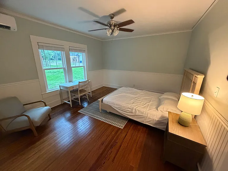 Furnished bedroom with hardwood floors, ceiling fan, large windows, and a dedicated desk area for working or studying.
