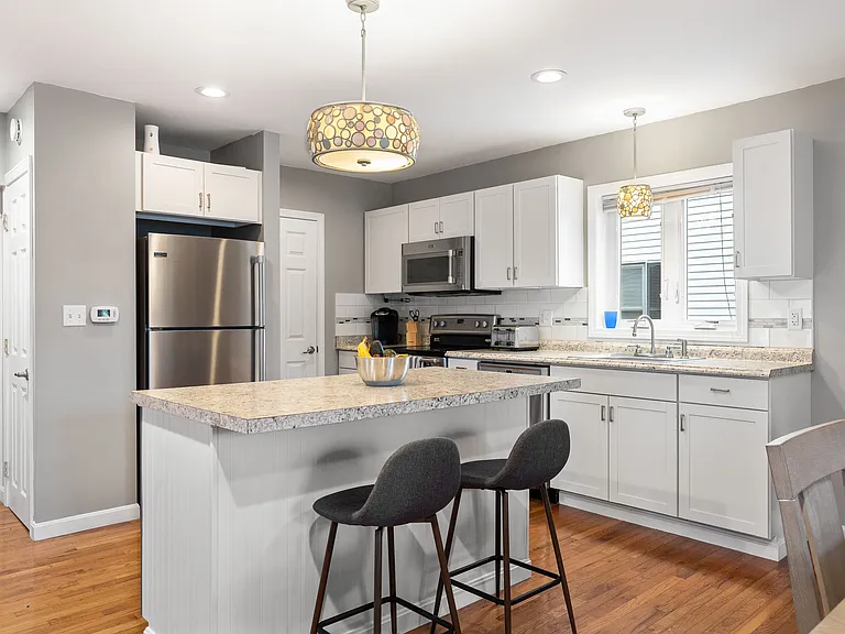 Stainless steel appliances, granite countertops, kitchen island, breakfast bar, pendant lighting, white cabinetry, hardwood flooring.