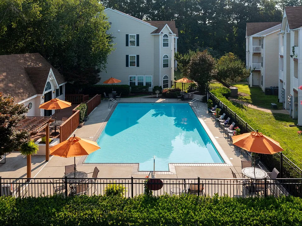 Resort-style community pool with turquoise water, orange umbrellas, and lounge chairs, framed by apartment buildings and lush trees at North Beach Apartments in Virginia Beach
