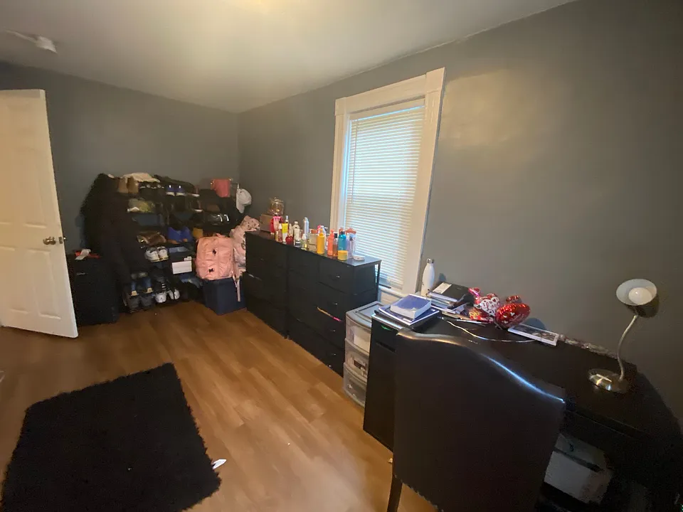 This room includes a large standing mirror, a work desk and chair, a work lamp, and a portable dresser, and shoe rack.