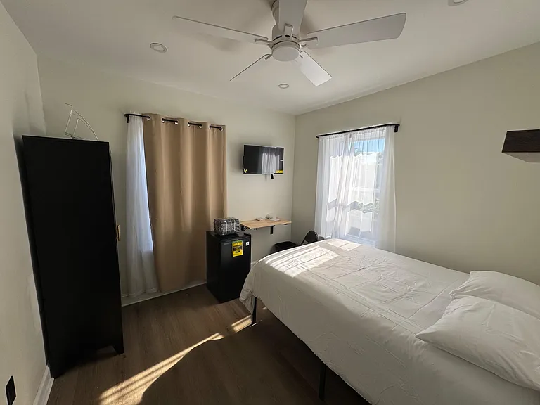 Private bedroom with ceiling fan, two windows with curtains, wall-mounted TV, wardrobe storage, and space for a full bed.