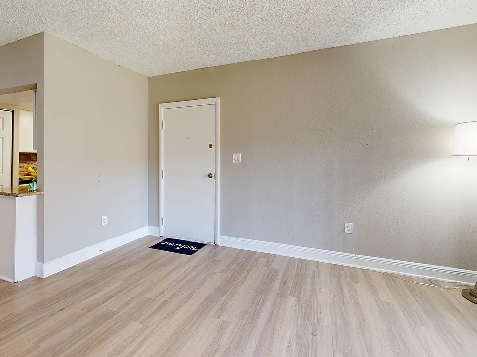 Inviting apartment interior with elegant wood flooring, modern floor lamp, and a glimpse into a cozy kitchen area.
