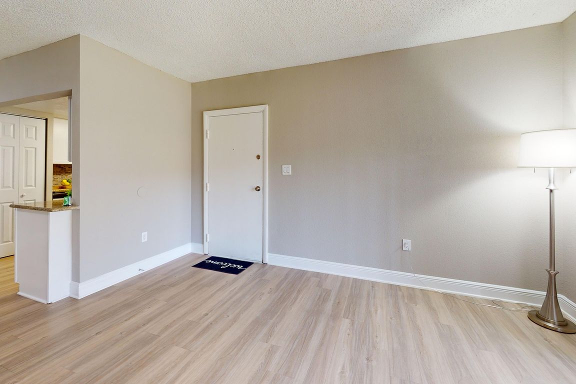 Inviting apartment interior with elegant wood flooring, modern floor lamp, and a glimpse into a cozy kitchen area.