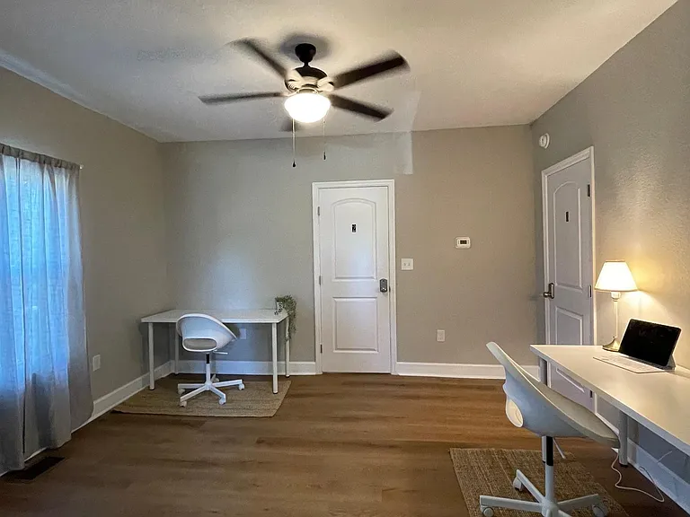 Spacious bedroom with wood-look floors, ceiling fan, and window with curtains, plus room for a desk or seating area.