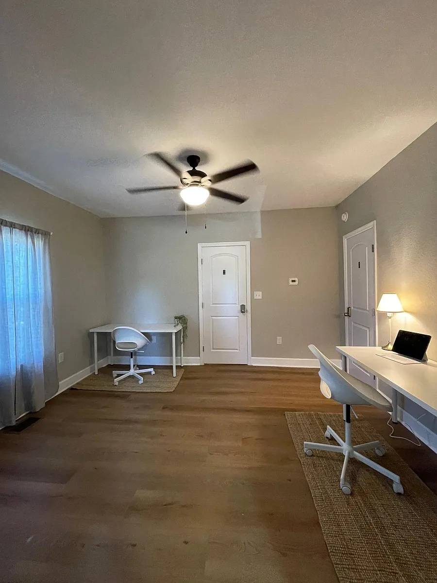 Spacious bedroom with wood-look floors, ceiling fan, and window with curtains, plus room for a desk or seating area. - Gilmore Ave