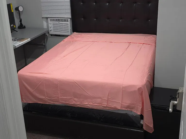 Private bedroom with a full-size bed, desk workspace, window blinds, and in-room window A/C for personalized comfort.
