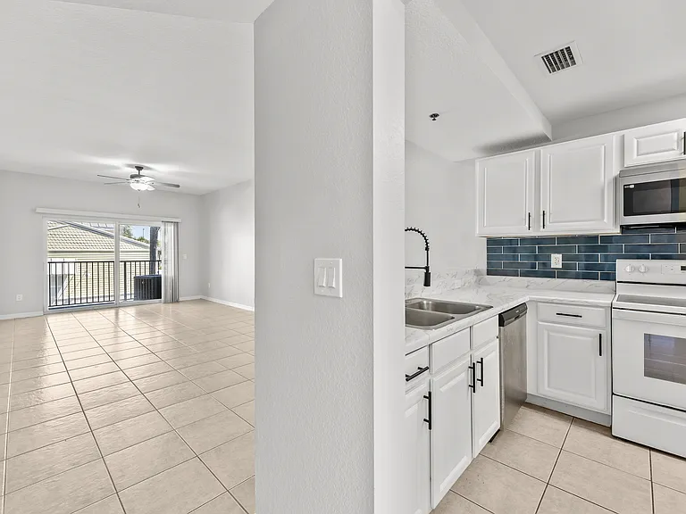 Modern open-concept living space featuring a sleek kitchen with white cabinetry, elegant blue backsplash, and spacious tile flooring leading to a bright living room with balcony access.