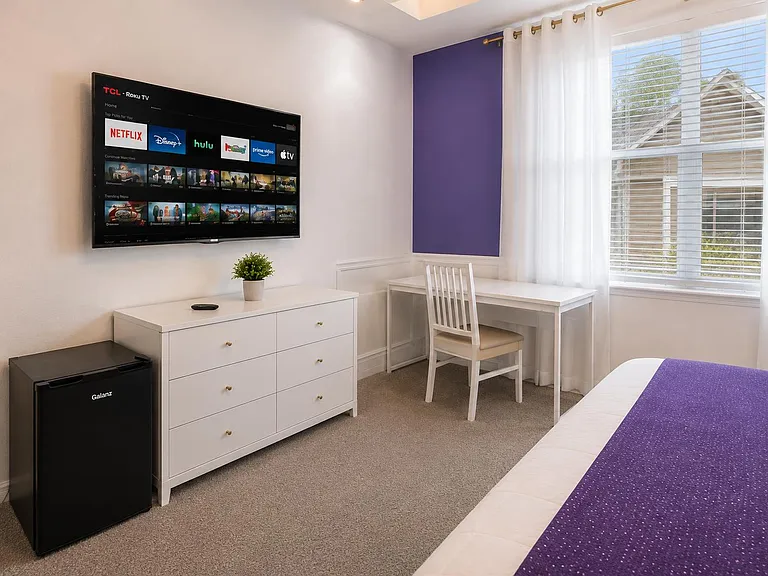 Private bedroom setup with a wall-mounted TV, dresser storage, mini fridge, and dedicated desk by a large window.