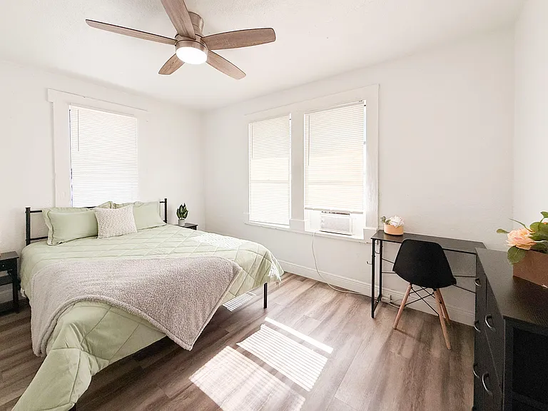 Furnished bedroom with a full bed, ceiling fan, large windows with blinds, and a dedicated desk/work area.