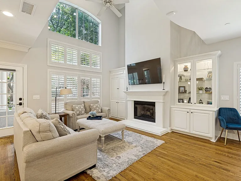 High-ceiling living area featuring extensive window treatments, a gas fireplace with a classic white mantel, built-in cabinetry with glass display shelves, a ceiling fan, and wood-finish flooring