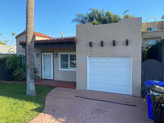 Charming Spanish-style house with mature palm trees, grassy lawn, and a brick pavestone driveway.