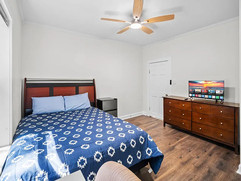 Furnished bedroom with a full bed, dresser with TV, mini fridge, and ceiling fan, plus a large window for natural light.