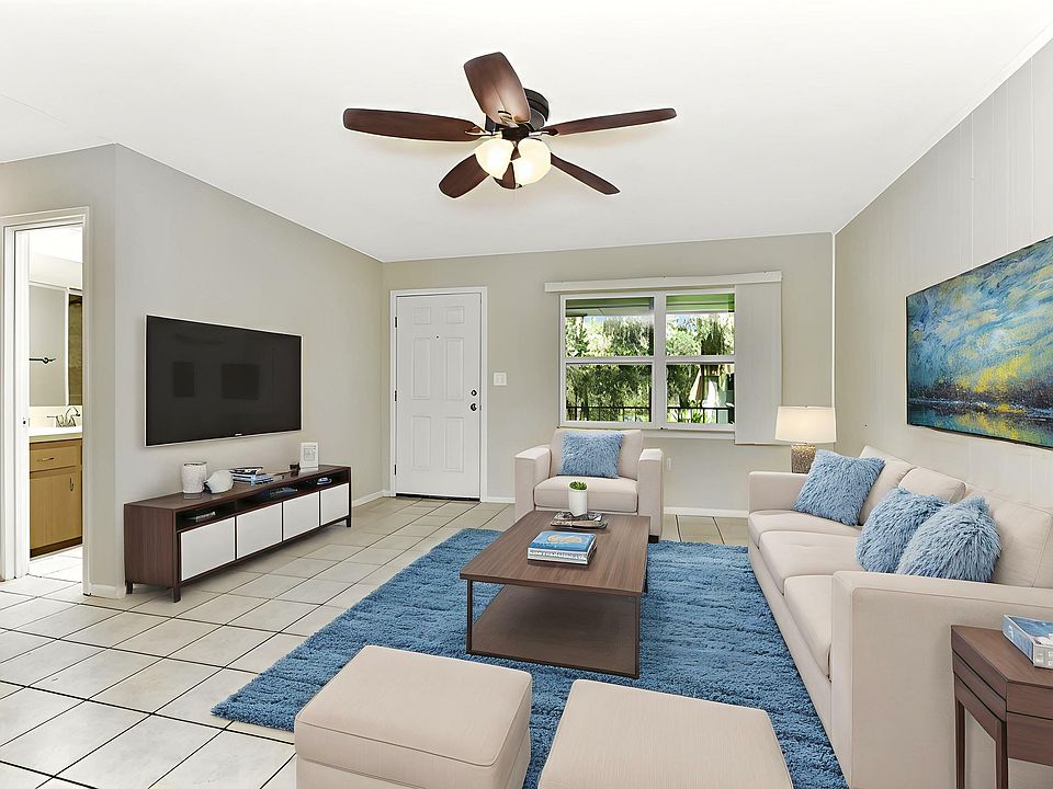 Bright and inviting living room with cozy white sofas, vibrant blue accents, and a stylish ceiling fan, complemented by a large window offering lush green views.