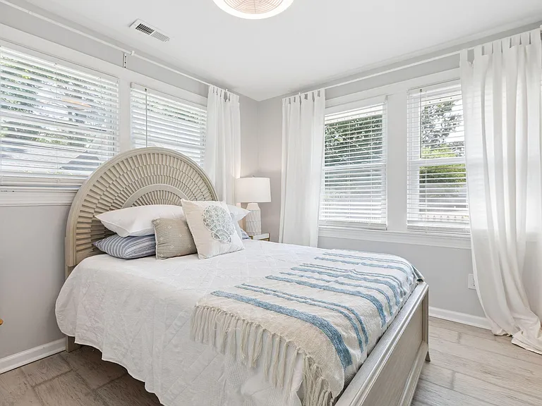 Sunlit bedroom with a comfortable bed, corner windows with blinds and curtains, and ample space for a nightstand.
