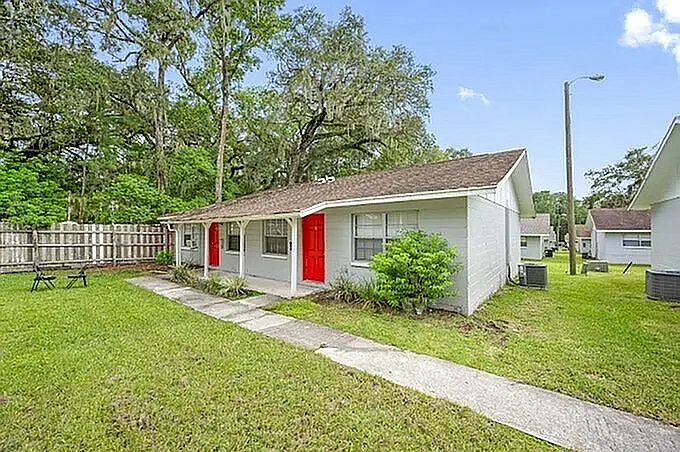Charming duplex with vibrant red doors, surrounded by lush greenery and mature trees, offering a cozy and inviting exterior.