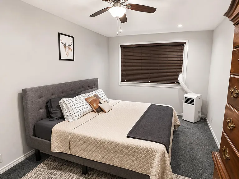Furnished bedroom with a queen bed, ceiling fan, and window blinds, plus dresser storage and portable A/C unit.