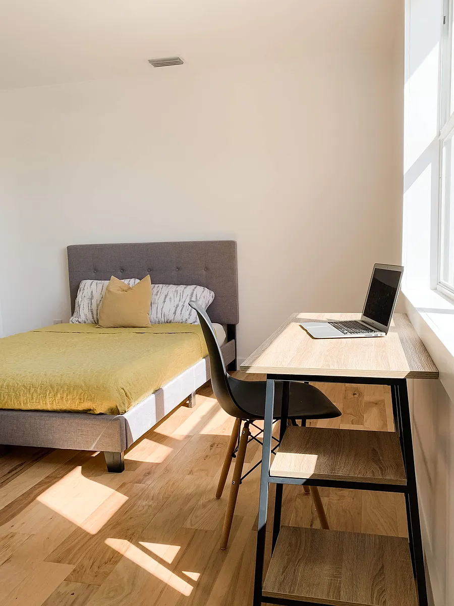 Furnished bedroom with a comfortable bed, dedicated desk workspace, and large window providing ample natural light. - 9th Ave N