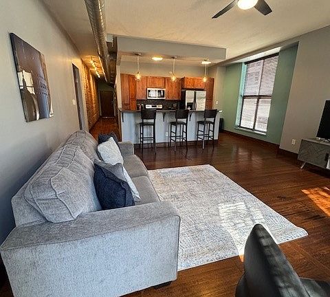 Stylish open-concept living area with a gray sofa, cozy rug, and sleek black chair. The kitchen features warm wood cabinets, stainless steel appliances, and a countertop with barstools. Dark wood floors and exposed ductwork add an industrial-modern vibe.