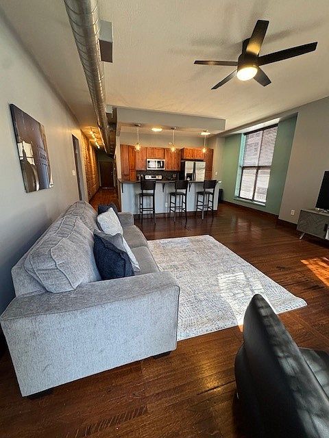 Stylish open-concept living area with a gray sofa, cozy rug, and sleek black chair. The kitchen features warm wood cabinets, stainless steel appliances, and a countertop with barstools. Dark wood floors and exposed ductwork add an industrial-modern vibe.