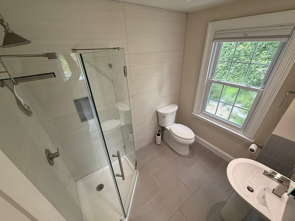 This renovated bathroom features a frameless glass shower, sleek tile flooring, a modern pedestal sink, and an LED mirror cabinet. Clean lines and quality finishes offer a stylish, space-efficient design perfect for everyday modern comfort.