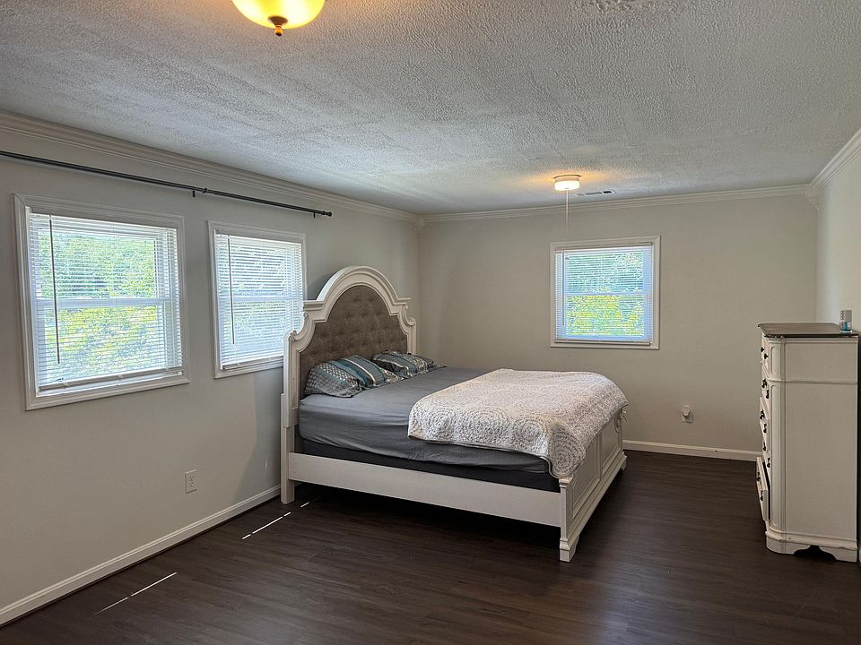 Spacious bedroom with three windows, a full bed, and a dresser, plus dark flooring and overhead lighting for easy living.