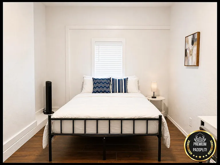 Clean bedroom with a full bed, nightstand lighting, and a window with blinds, plus space for a desk or dresser.