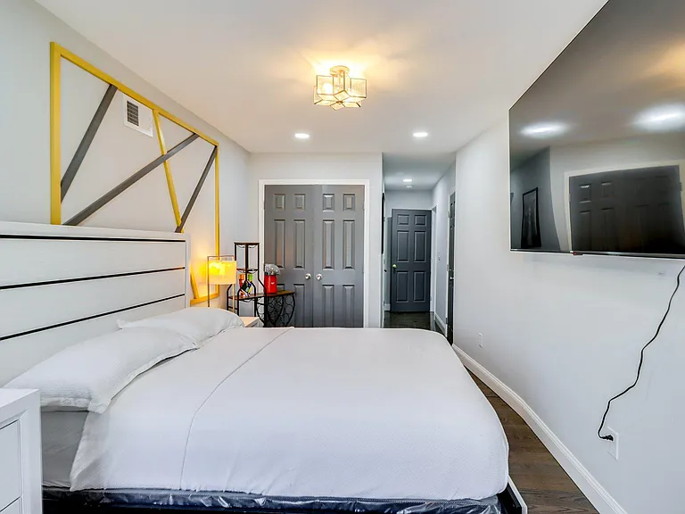 Furnished bedroom featuring a full-size bed, bedside lighting, wall-mounted TV, and double-door closet for storage.