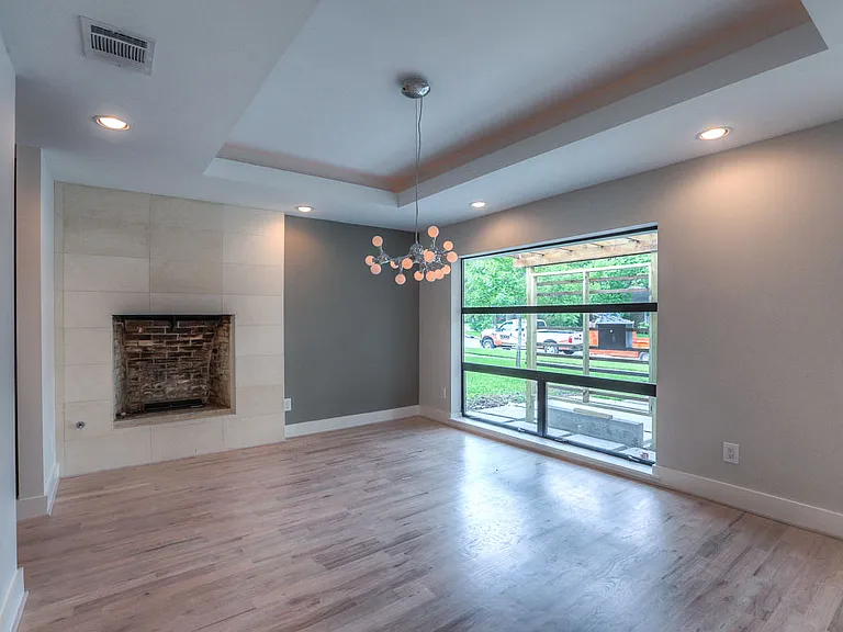 Bright dining room with a tile-clad fireplace, designer bubble chandelier, recessed tray ceiling, oversized picture window, and new hardwood floors installed during renovation.