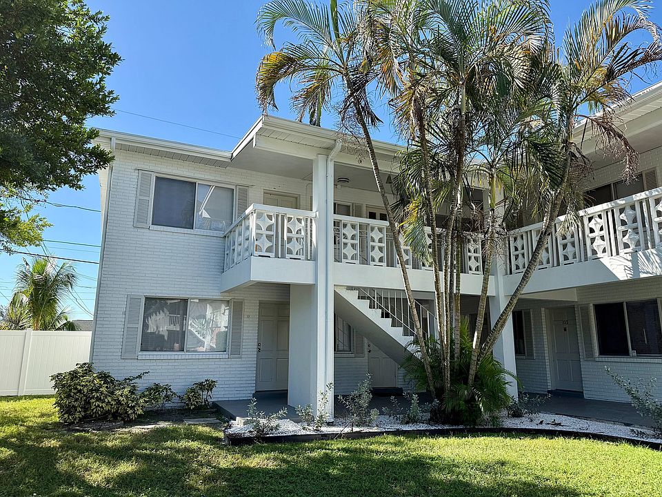 Charming two-story apartment building with white brick exterior, lush palm trees, and inviting balconies under a clear blue sky.