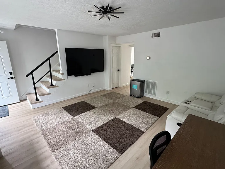 Open living area with wood-look flooring, ceiling fan, wall-mounted TV, and seating space near the staircase.