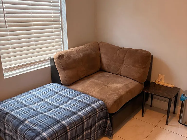 Compact sleeping area with a daybed-style setup, side table, and window with blinds for light and privacy.