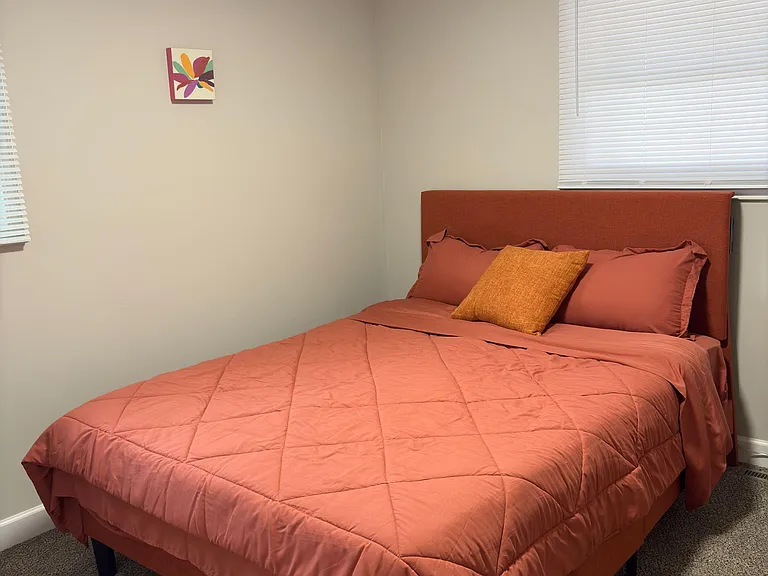 Furnished bedroom featuring a comfortable bed, soft carpeting, and a window with blinds for light and privacy.