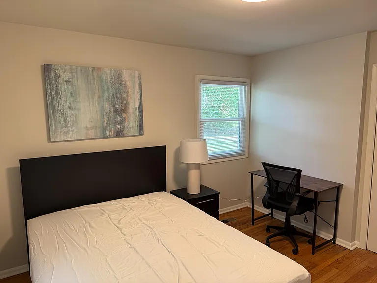 Furnished bedroom with a full bed, nightstand lamp, and a dedicated desk setup near a window with blinds.