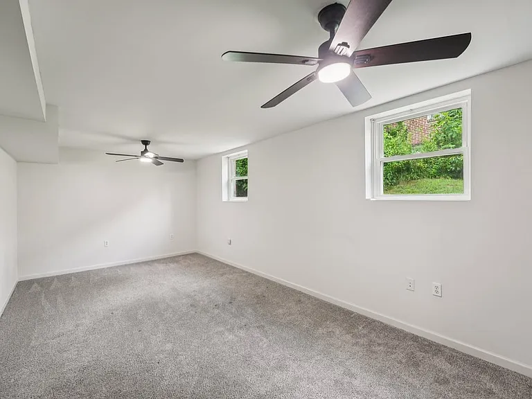 A spacious lower level room with soft carpeting, crisp white walls, and modern ceiling fans, creating a flexible retreat for lounging, resting, or quiet everyday escape.