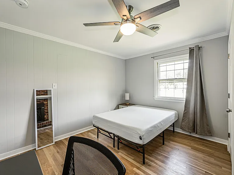 Private bedroom with a ceiling fan, hardwood-style floors, and a large window with curtains; includes a nightstand and space