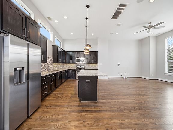 Open-concept kitchen and living area with dark wood cabinetry, stainless steel appliances, and a large island with pendant lighting. High ceilings, hardwood floors, and abundant natural light create a bright, inviting space.