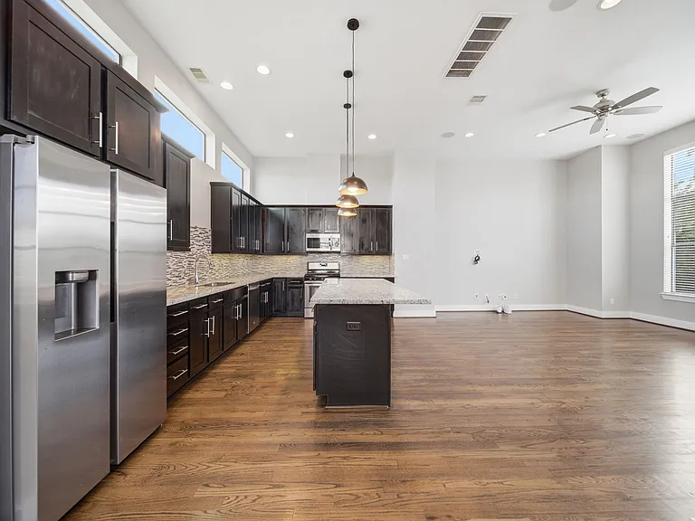 Open-concept kitchen and living area with dark wood cabinetry, stainless steel appliances, and a large island with pendant lighting. High ceilings, hardwood floors, and abundant natural light create a bright, inviting space.