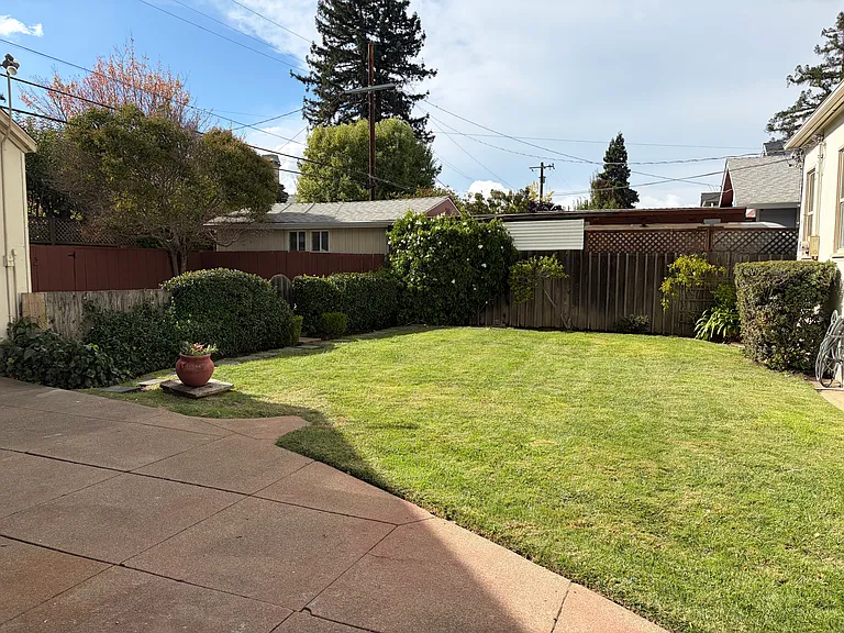 Large, level grassy yard with a patio area behind the low hedges to the left.