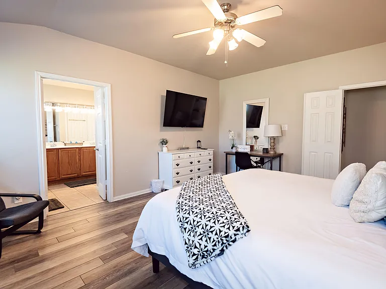 Furnished bedroom with ensuite bath access, ceiling fan, wall-mounted TV, and a dedicated desk area for work or study.