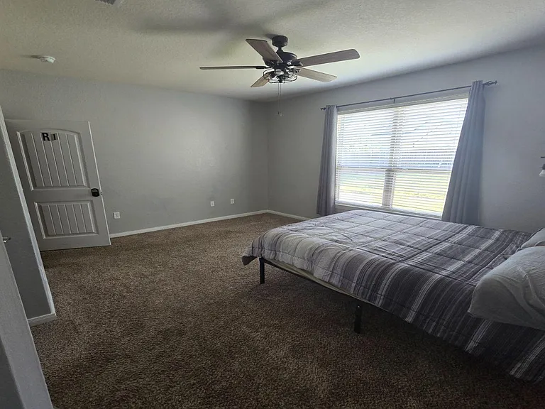 Carpeted bedroom with a large window, ceiling fan, and space for a full/queen bed plus a small desk or dresser.