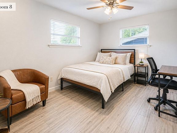 Furnished bedroom with a full bed, two windows, ceiling fan, nightstand, and a dedicated desk workspace plus lounge chair.