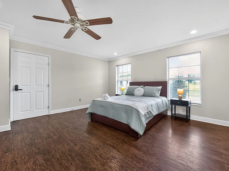 Spacious bedroom featuring hardwood floors, ceiling fan, and large windows with blinds, plus room for nightstands.