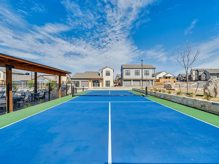 A tennis court with a blue surface and white lines is surrounded by houses and trees.