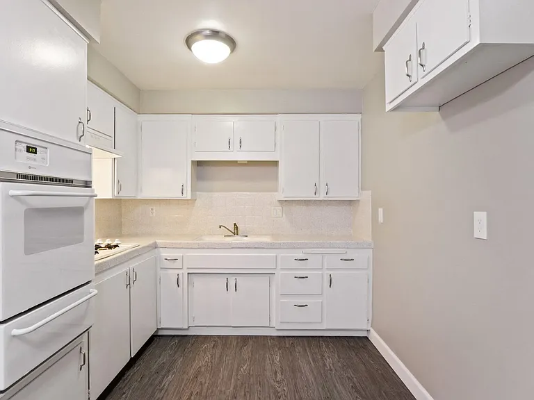Kitchen with White oven, cooktops on the left, double white kitchen sink, white kitchen cabinets and cupboards, Tile backsplash and countertops, wood style flooring