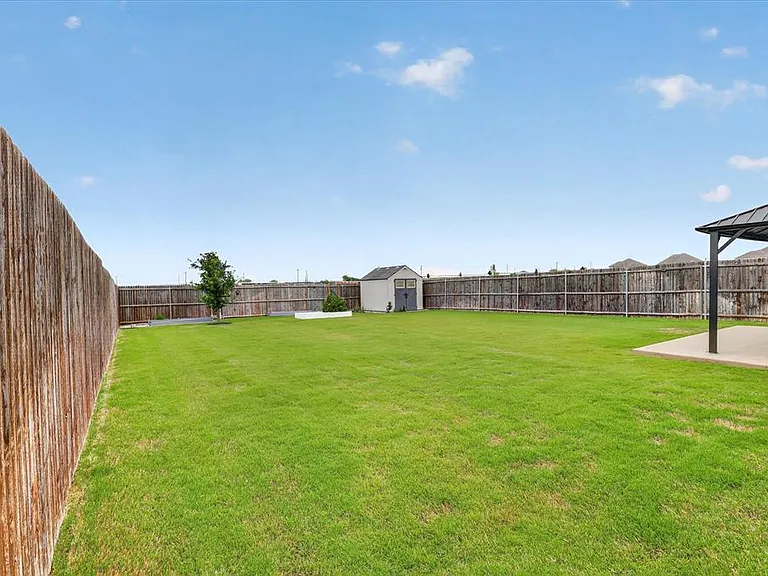 Expansive backyard featuring a lush lawn, privacy fencing, a freestanding storage shed, and a covered patio area