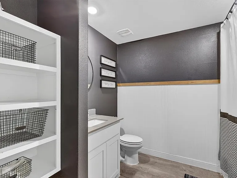 Bathroom featuring a built-in white shelving unit, a white vanity with an undermount sink, a toilet, a full shower with a white curtain, and wood-finish flooring
