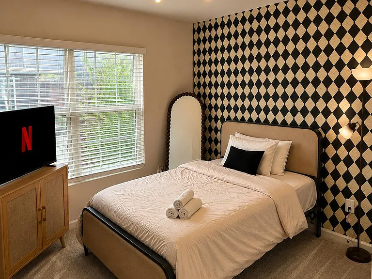 Furnished bedroom with a full bed, accent wall, dresser, and large window with blinds for natural light.